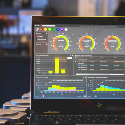 Nærbilde av en bærbar datamaskin som viser et ERP-dashbord med grafer, målere og analyser for produksjonsdata. Skjermen inneholder KPI-indikatorer og statistikk, mens bakgrunnen er uskarp med industrielt utstyr.