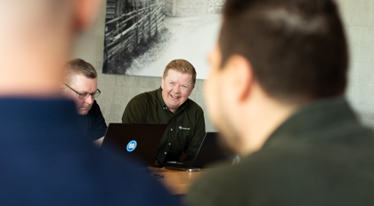 A group of men sitting around a laptop computer with a Monitor ERP logo. The men are happy and are smiling.