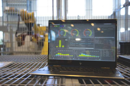 Laptop displaying industrial monitoring software with colorful graphs and gauges, set on a metal surface with industrial equipment in the background