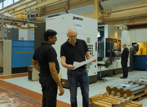 To menn i svart arbeidstøy står i en industrihall og ser på et dokument. I bakgrunnen er en stor maskin med teksten ‘Okuma’, samt hyller fylt med utstyr og en medarbeider som jobber ved en maskin. Flere metallstenger ligger stablet på paller i forgrunnen.