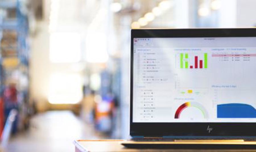 En bærbar PC med et dashbord som viser grafer og dataanalyse står på et bord i et industrielt lager- eller produksjonsmiljø. Fargerike diagrammer og grafer vises på skjermen, mens bakgrunnen er uskarp med reoler og utstyr.