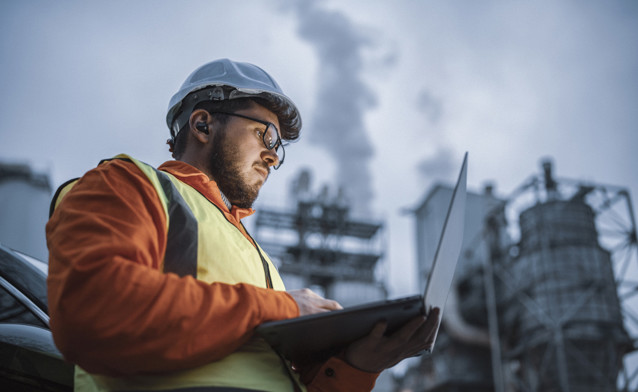 Industriarbeider iført vernehjelm, refleksvest og vernebriller bruker en bærbar datamaskin på et olje- eller gassanlegg. Fabrikkstrukturer og røykskyer sees i bakgrunnen, som illustrerer overvåkning og digitalisering i tungindustrien.